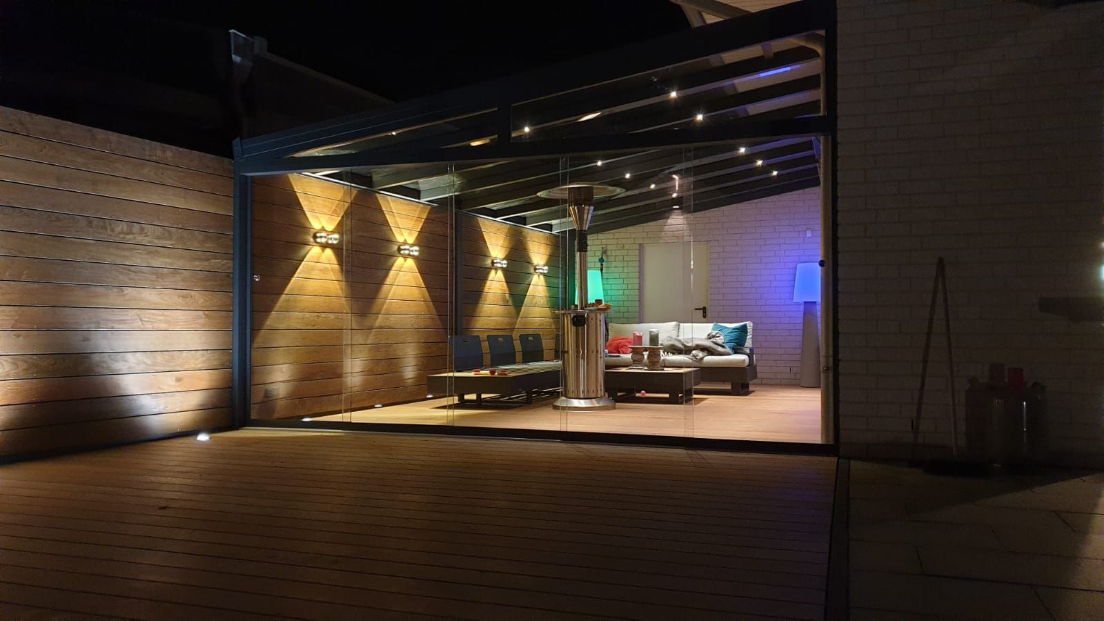 Ein moderner Lounge-Bereich im Freien bei Nacht, mit einer holzgetäfelten Wand, warmen Wandleuchten, einem Glasdach und Möbeln.