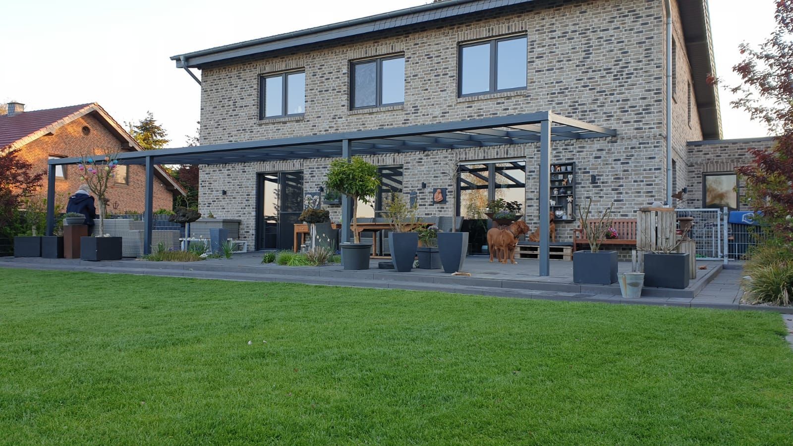 Ein Backsteinhaus mit einer dunklen Metall-Terrassenüberdachung über einer gepflasterten Terrasse, die auf einen gepflegten grünen Rasen bei Sonnenuntergang blickt.