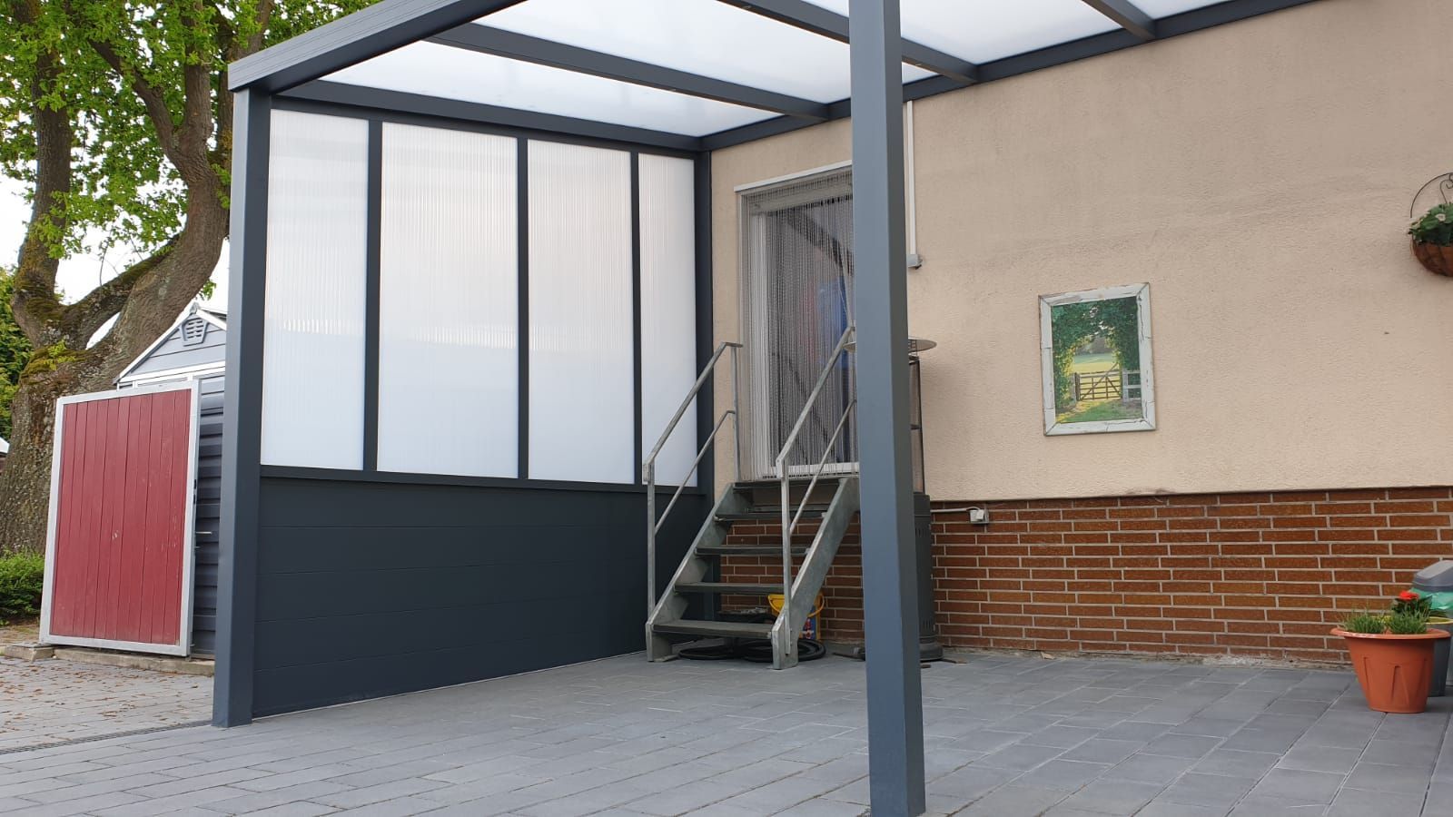 Eine Terrasse mit modernem grauen Dach und Glasseitenwand, Metalltreppen, die zu einer Haustür führen, und ein Ziegelsteinfundament.
