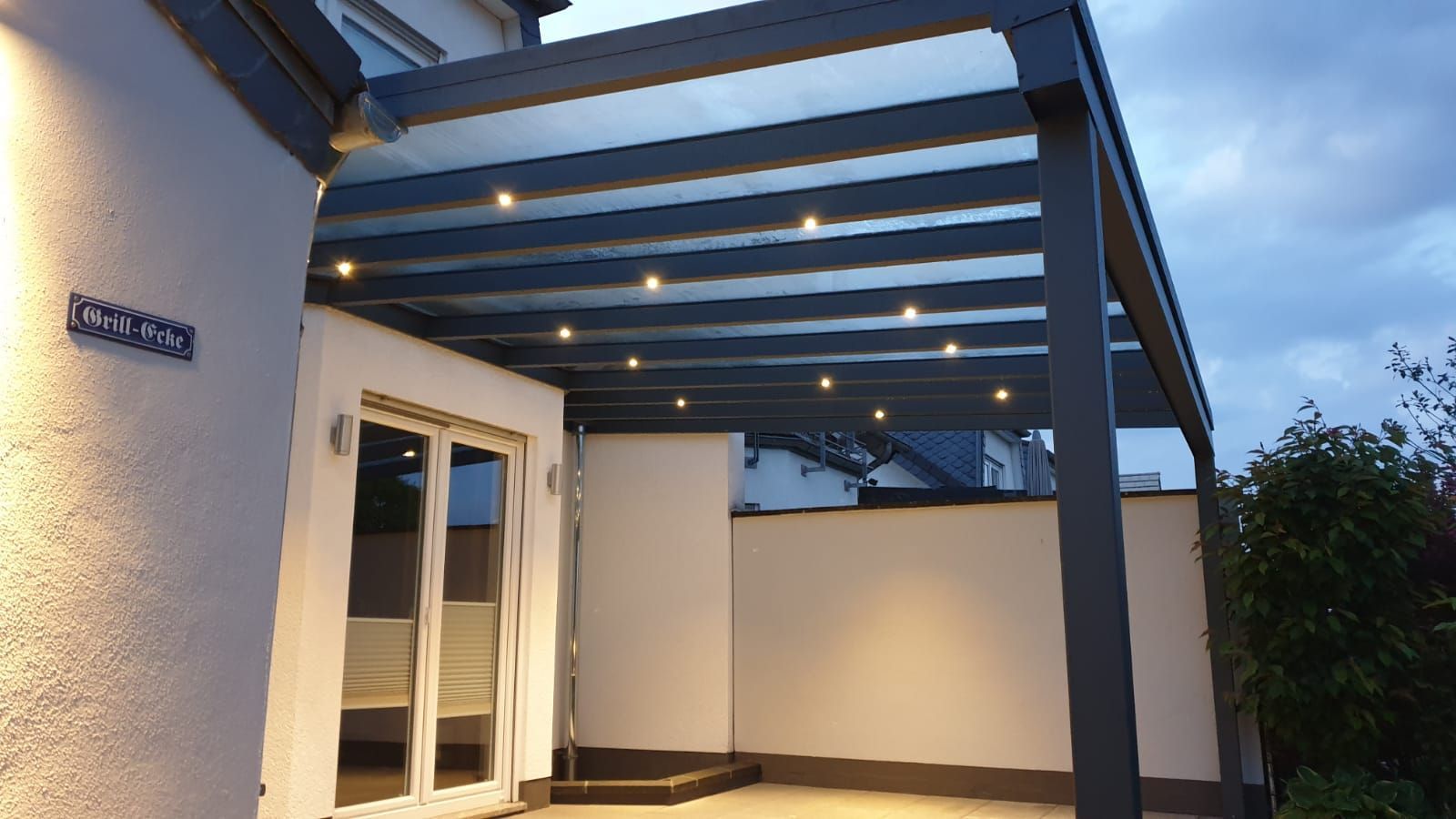 Eine graue, moderne Terrassenpergola, die an ein weißes Haus angebaut ist, mit integrierter Einbaubeleuchtung, die eine gepflasterte Terrasse beleuchtet.