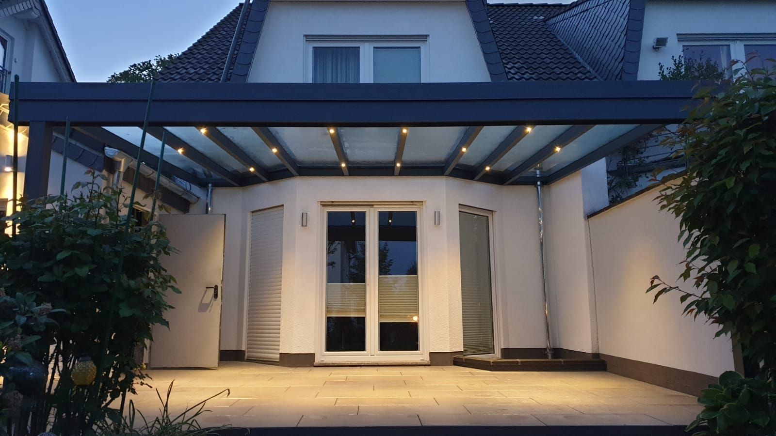 Eine beleuchtete Terrasse mit einem gläsernen Dach, die an ein weißes Haus angebaut ist, in der Abenddämmerung, mit Steinboden und Grünpflanzen.