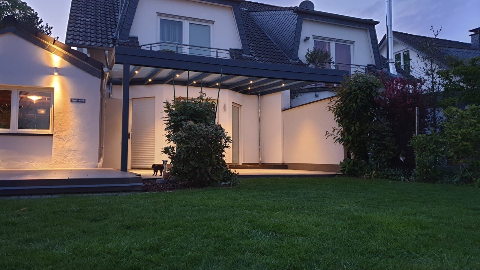 Eine Terrasse mit einer dunklen, modernen Pergola und integrierter Beleuchtung, die an ein zweistöckiges weißes Haus angrenzt, in der Abenddämmerung.