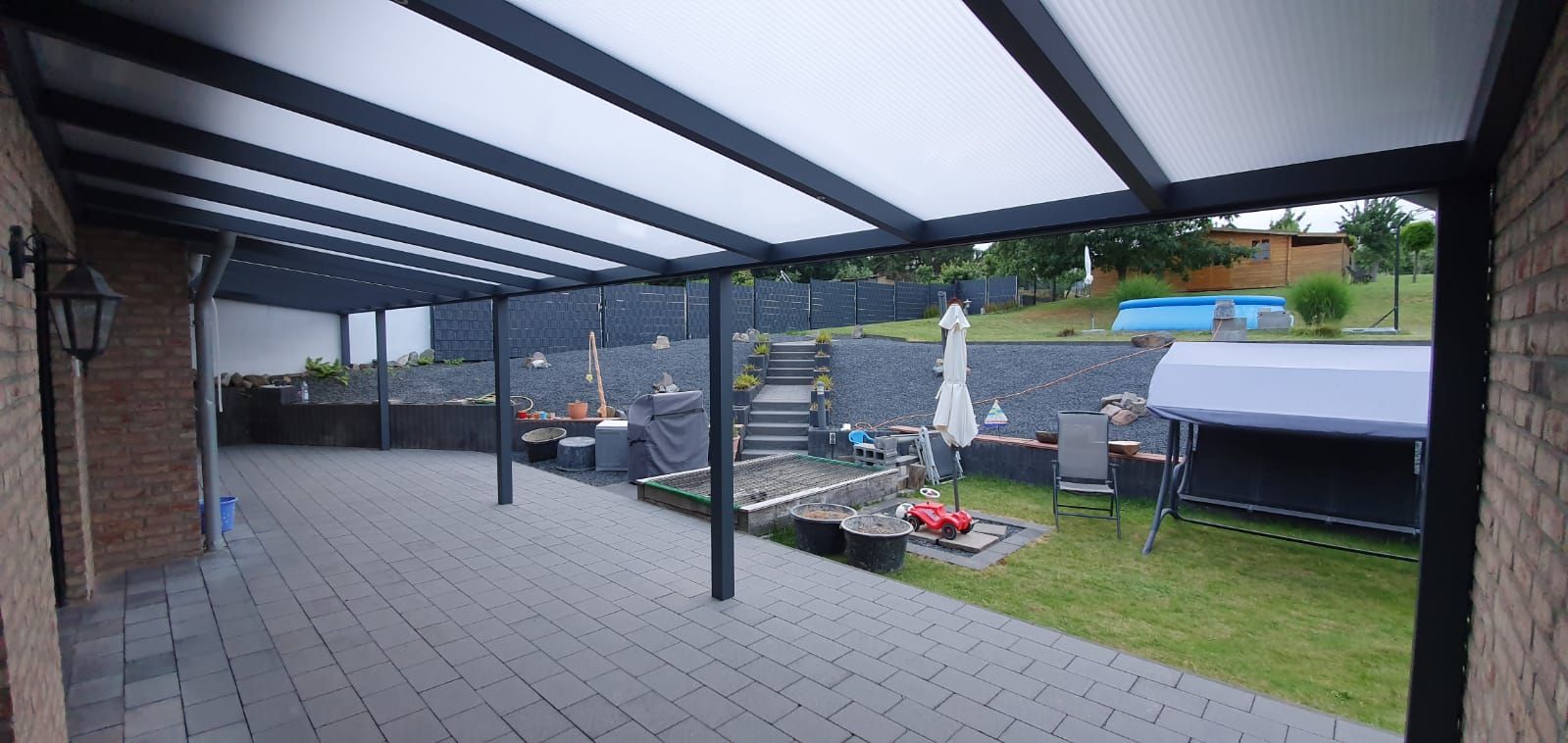 Eine Terrasse mit einem dunkelgrauen Pergola-Dach aus Metall, die auf einen Hinterhof mit gepflasterten Stufen, Rasen und einem oberirdischen Pool blickt.