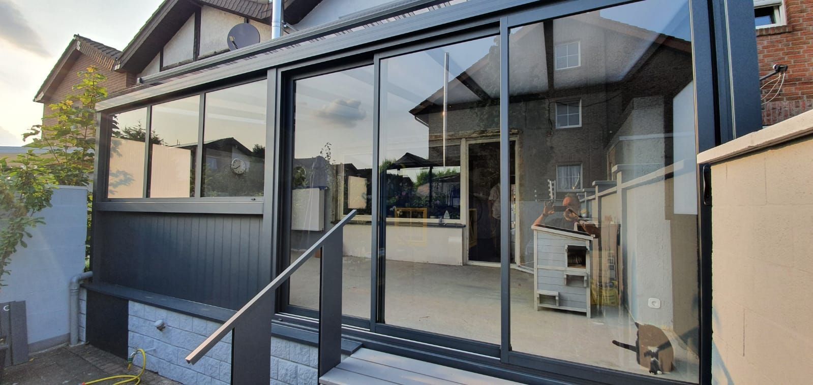 Ein moderner, dunkelgrau gerahmter Glaswintergartenanbau, der an ein Backsteinhaus angebaut ist und über eine kleine Treppe und ein Metallgeländer verfügt.