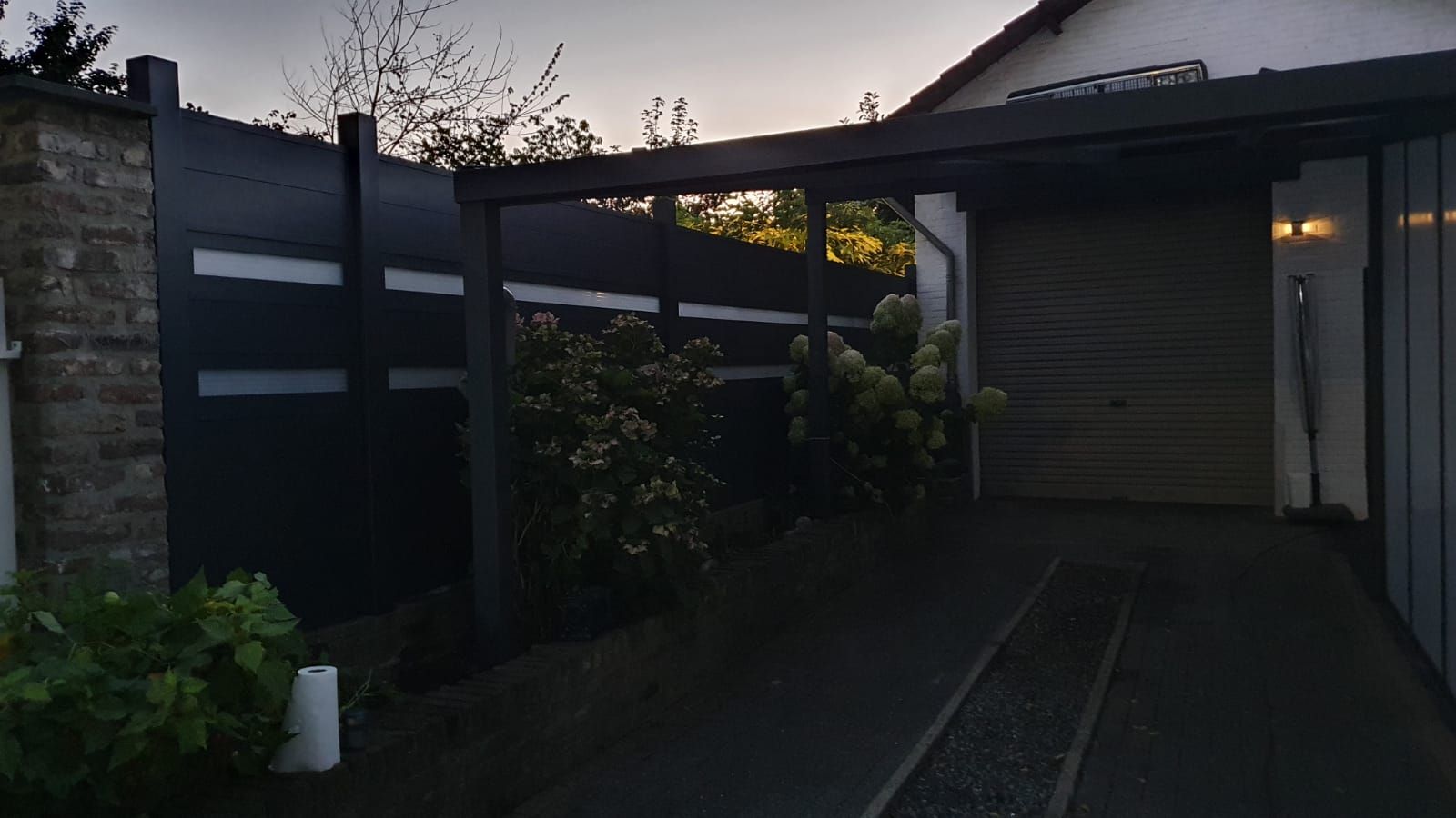 Ein moderner, dunkelgrauer Zaun mit horizontalen Lichtstreifen neben einer Einfahrt und einer Garage in der Abenddämmerung.