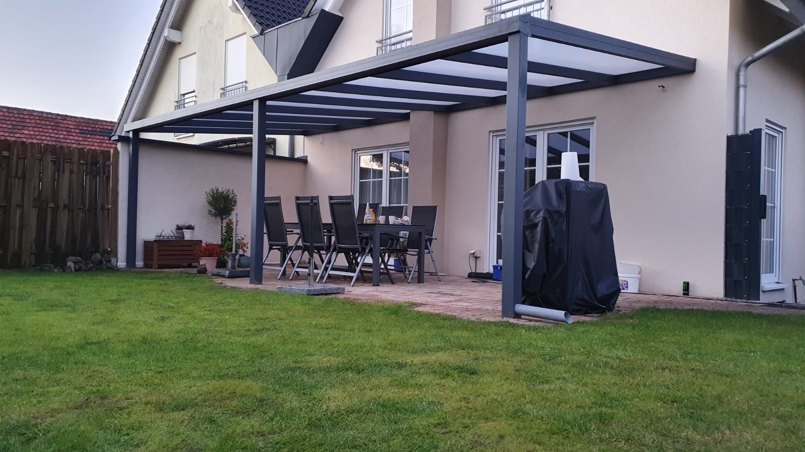 Eine moderne, dunkelgraue Terrassenüberdachung, die an ein beiges Haus angebaut ist, mit Gartenmöbeln und einem Grill auf einer Steinterrasse.