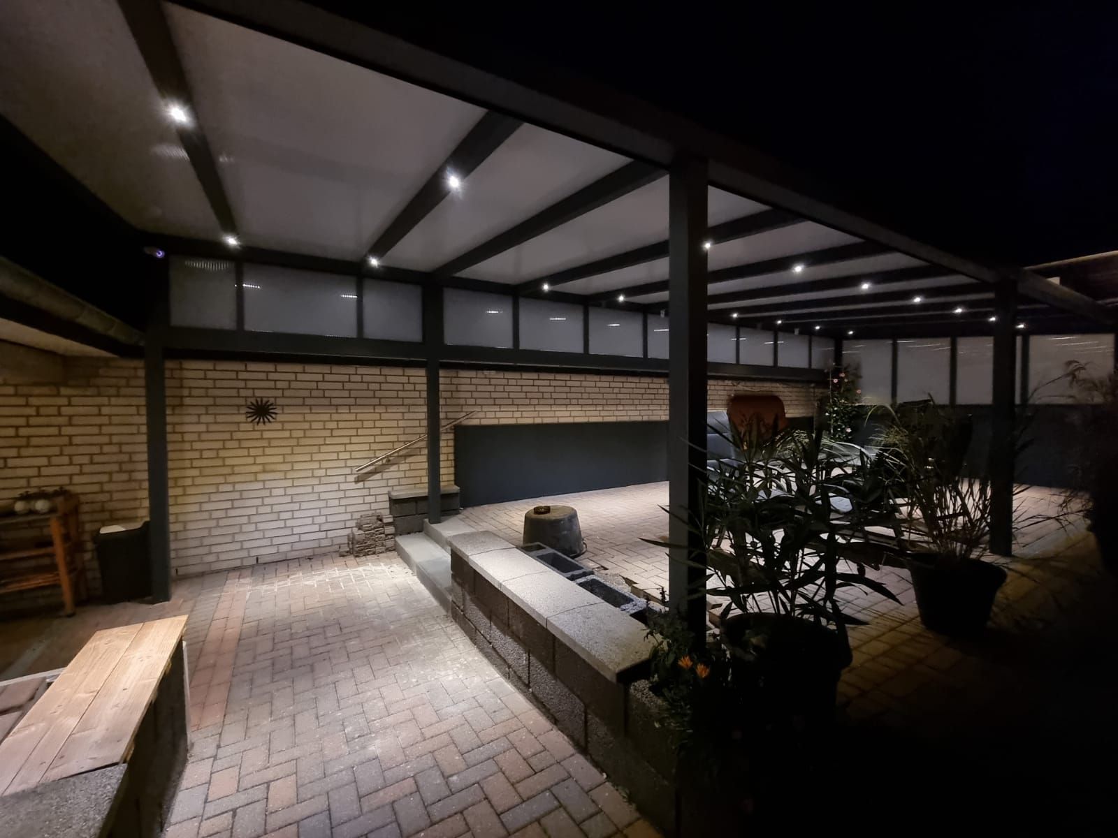 Eine dunkle, moderne Terrasse bei Nacht mit überdachter Holzpergola, Steinboden, Backsteinmauer und sanfter Beleuchtung.
