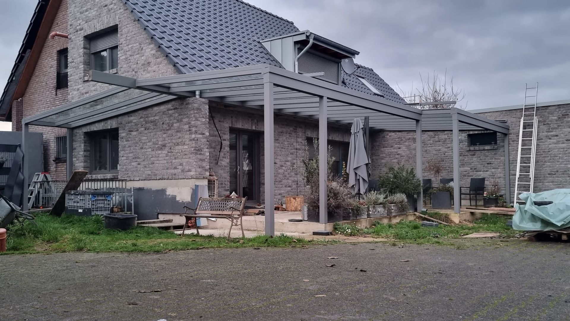 Ein von Steinmauern umgebenes Haus mit einer großen, grauen Metallpergola an der Rückseite und einem Kieshof davor.