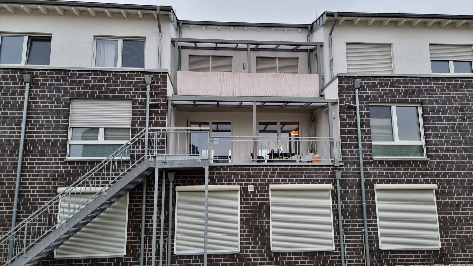 Ein mehrstöckiges Wohnhaus aus Backstein mit einer Außentreppe aus Metall, zwei zentralen Balkonen und Jalousien.