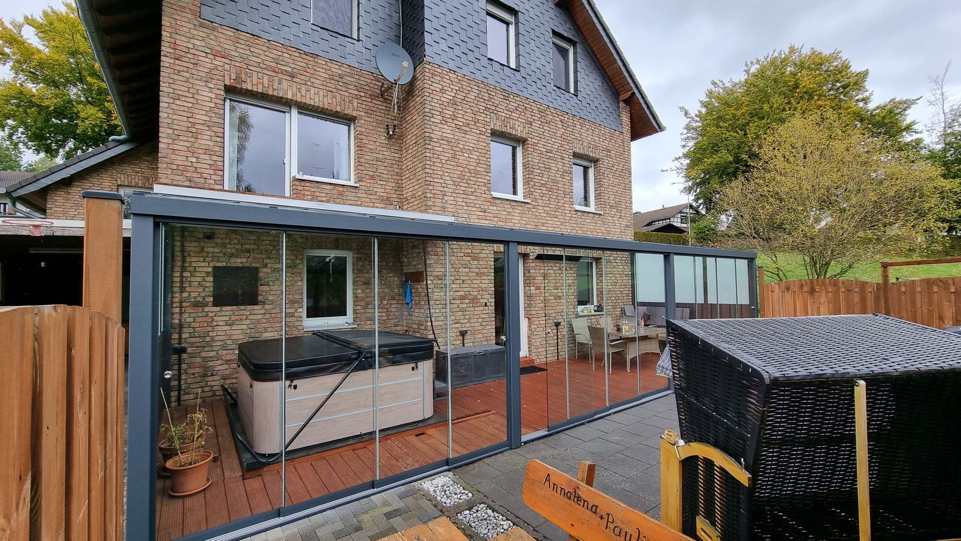 Eine Terrasse mit Whirlpool und Sitzgelegenheiten, umschlossen von einer verglasten Konstruktion, die an ein Backsteinhaus mit Schieferdach angebaut ist.