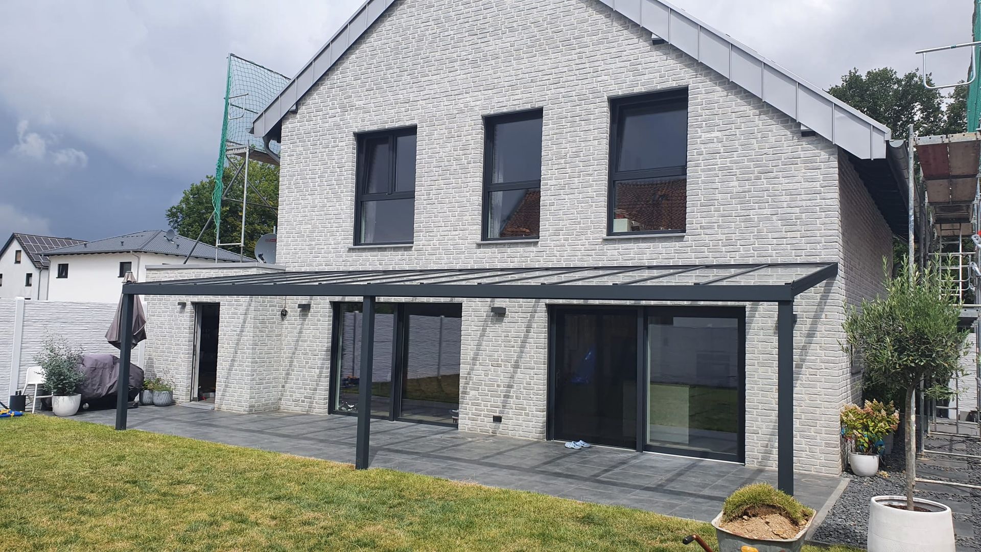 Ein modernes Haus mit strukturierten weißen Steinwänden, schwarzen Fensterrahmen und einer überdachten Terrasse mit Glasdach über einer gefliesten Terrasse.