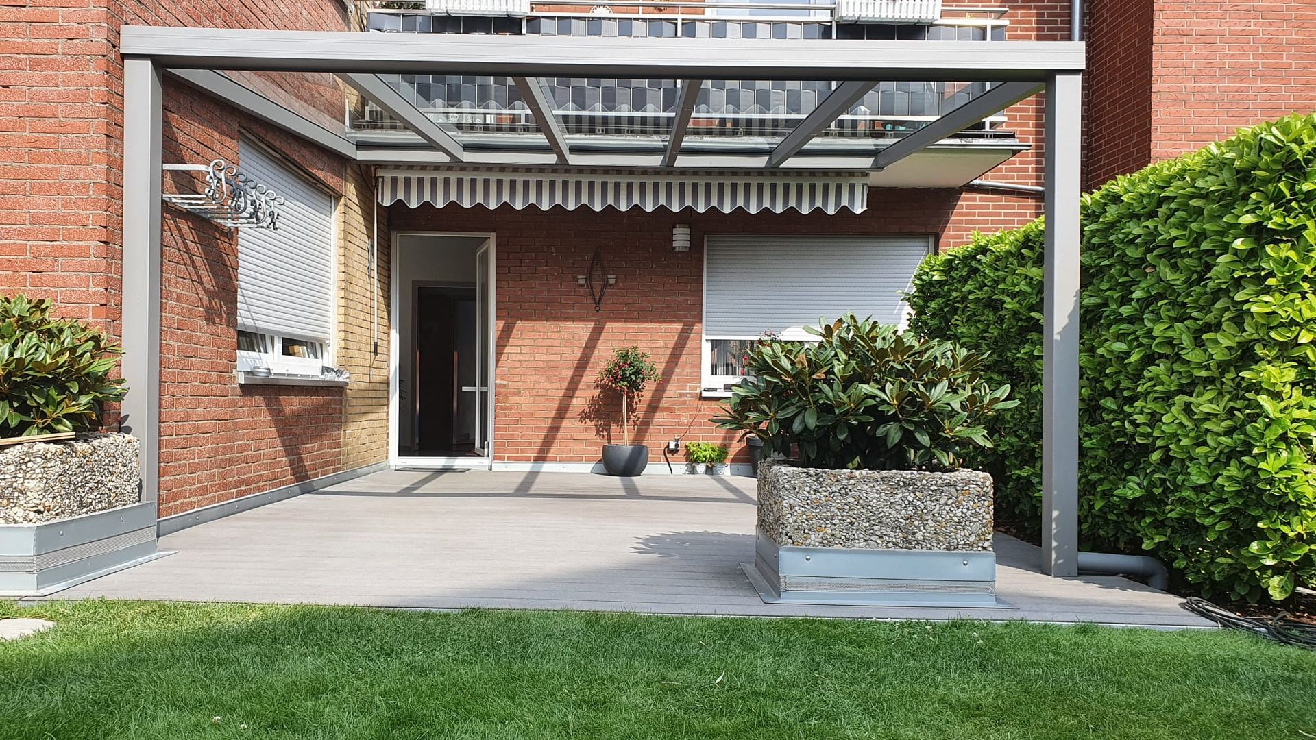 Die Terrasse eines Backsteinhauses verfügt über eine moderne graue Metallpergola, einen Steinpflanzkasten und eine Glasschiebetür, die ins Haus führt.