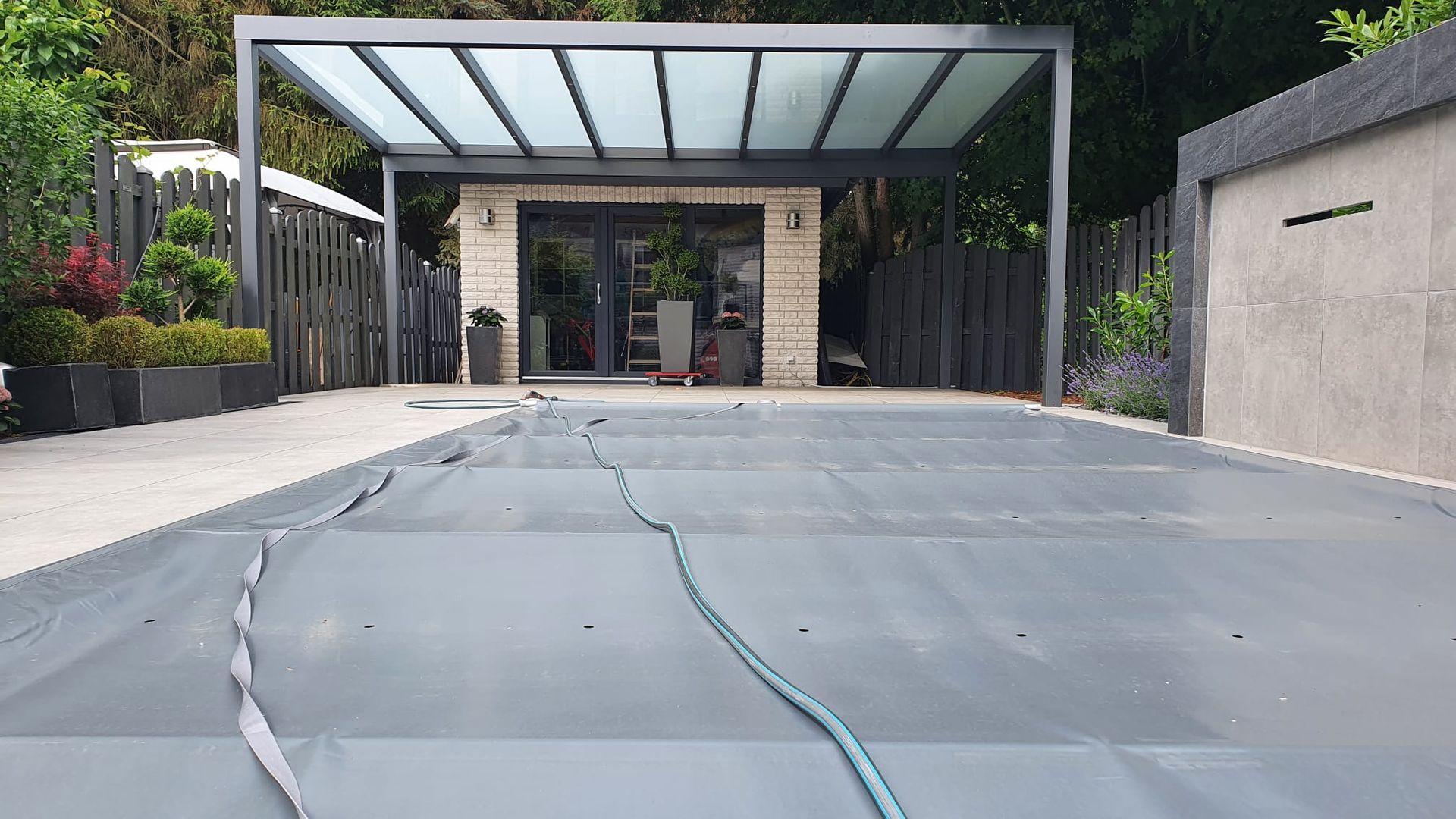 Auf einer Terrasse im Hinterhof befindet sich eine graue Poolabdeckung. Im Hintergrund sind eine moderne Außenanlage und eine Steinmauer zu sehen.