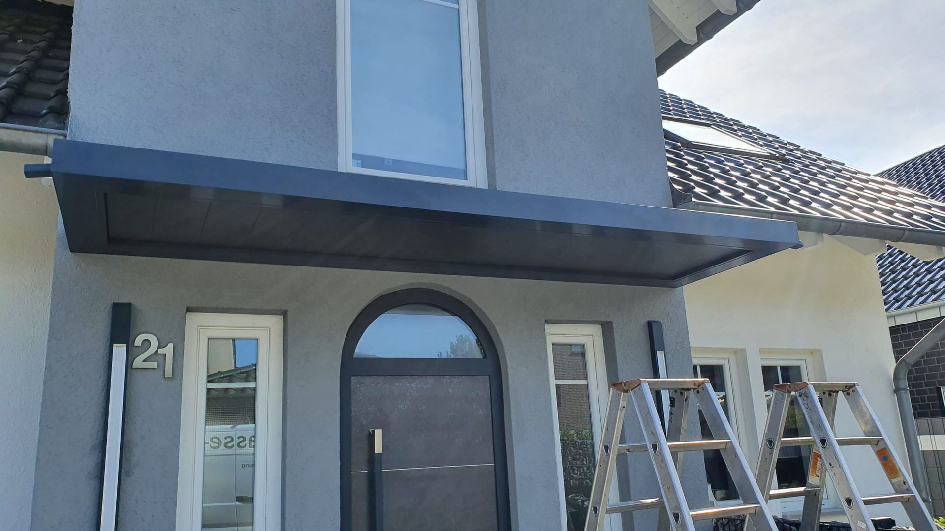 Ein grauer Hauseingang mit einem modernen, dunklen Vordach über der Haustür, flankiert von zwei Leitern auf der Auffahrt.