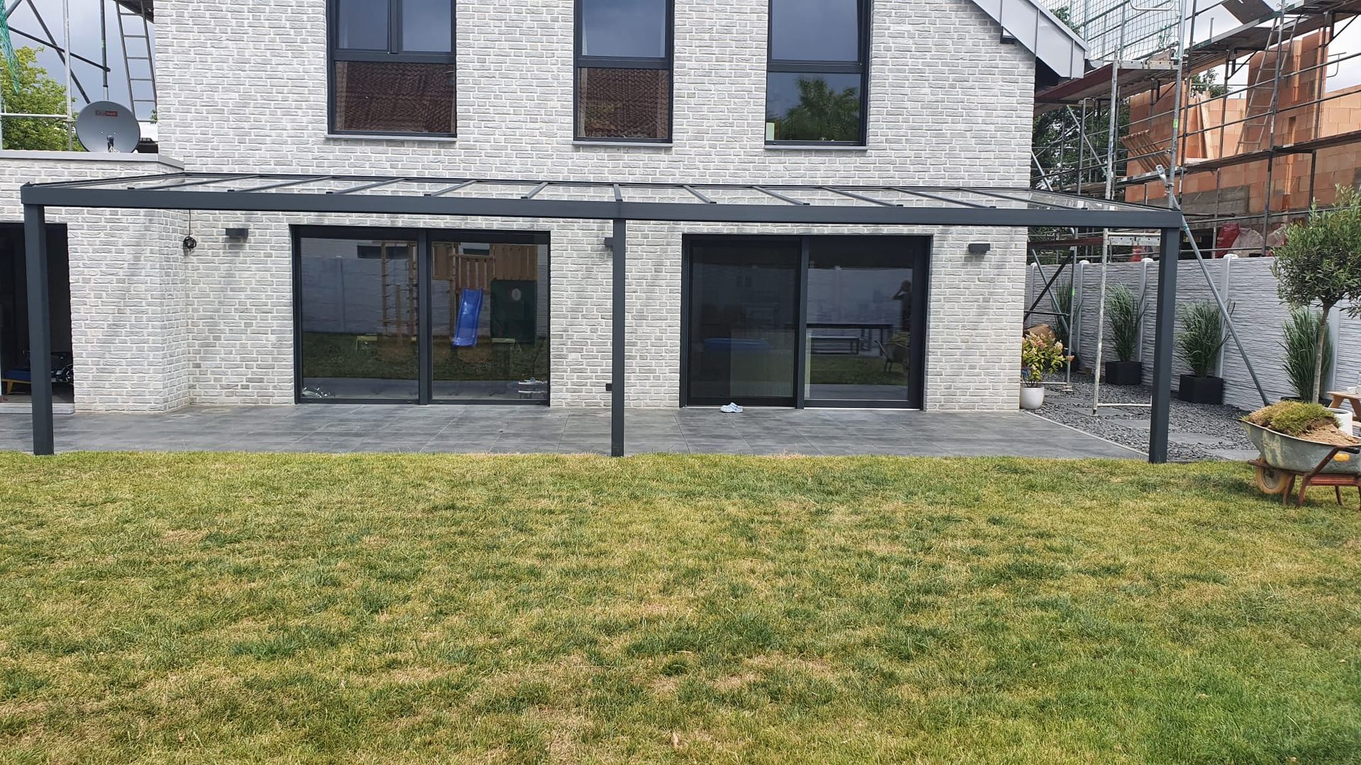 Ein Haus mit Steinfassade, Glasschiebetüren und einer modernen, dunkelgrauen Pergola auf einer Terrasse mit Blick auf einen Rasen.