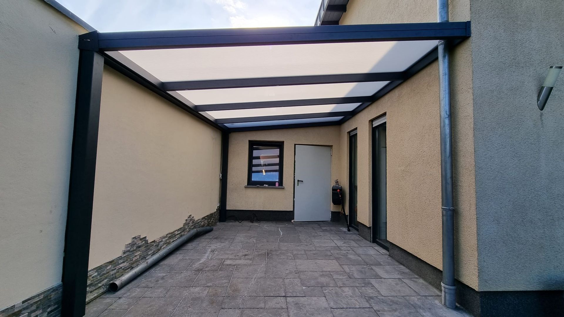 Eine von einem schwarzen Rahmen und einem lichtdurchlässigen Dach überdachte Terrassenfläche, die zwischen zwei beigen Außenwänden liegt.