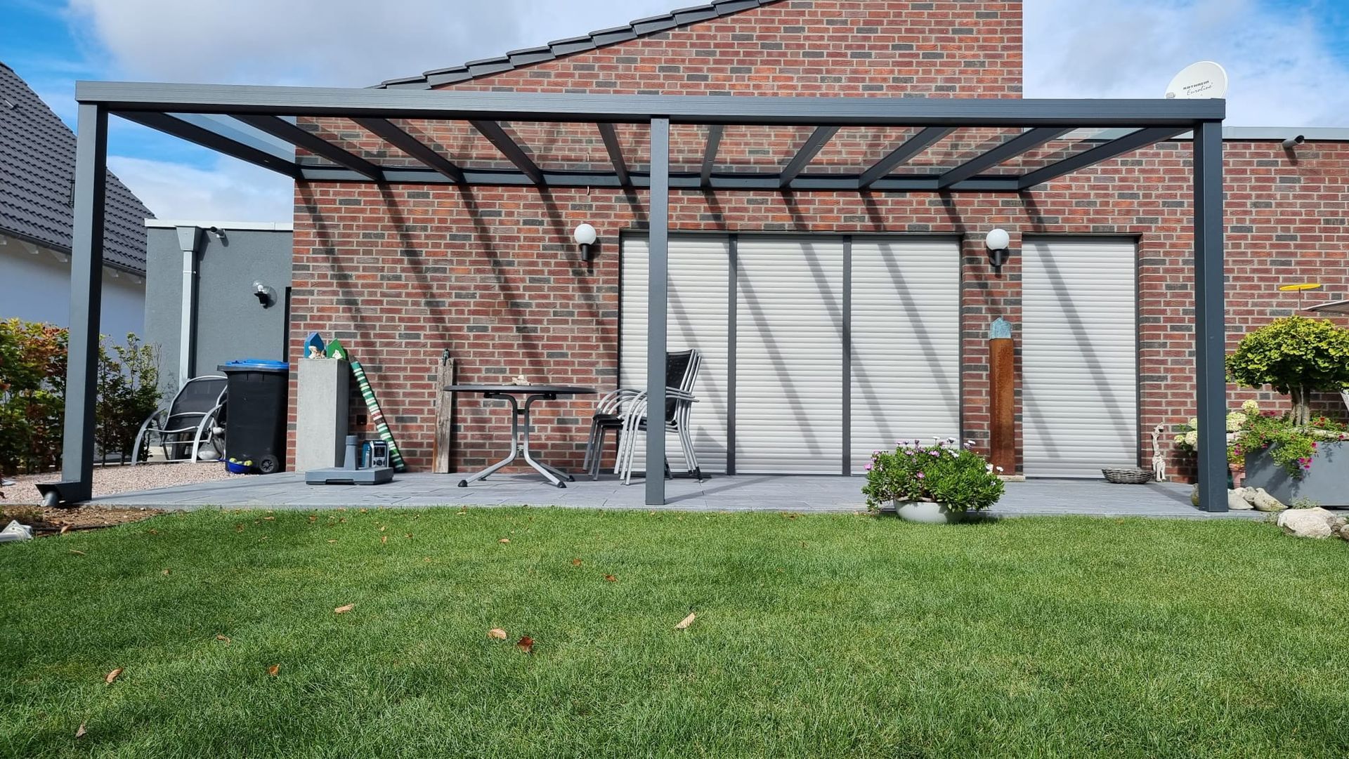 Eine graue Terrassenüberdachung aus Metall, die an einer roten Backsteinhausmauer befestigt ist und einen Blick auf einen grünen Rasen mit Tisch und Stühlen bietet.