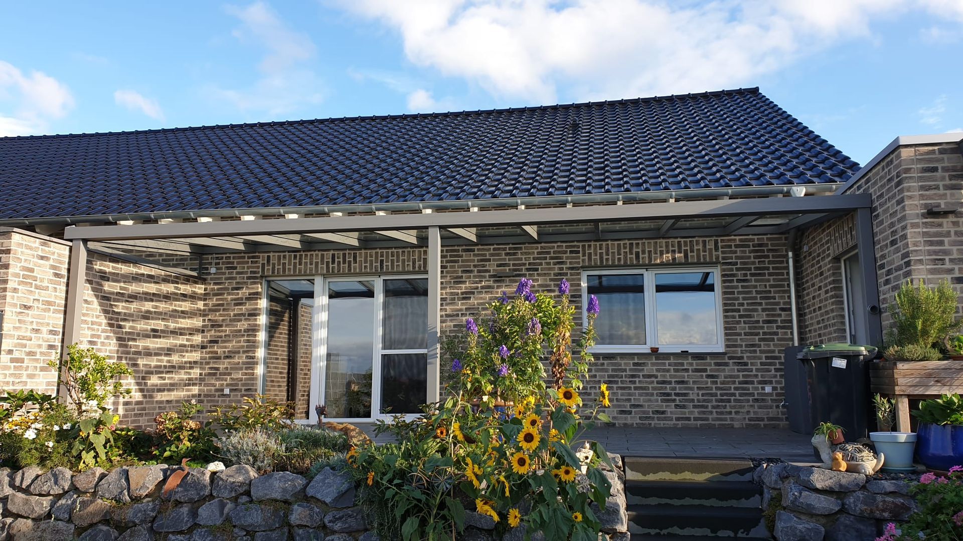 Ein Steinhaus mit dunklem Ziegeldach, einer hölzernen Pergola und einem davorliegenden Blumengarten unter sonnigem, blauem Himmel.