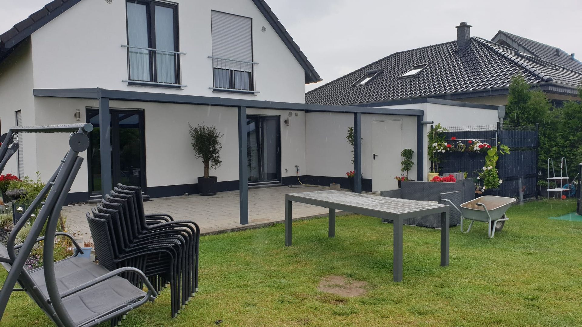 Ein weißes Haus mit grauer Terrassenüberdachung, Gartenmöbeln, einem Tisch und einem Hängesessel in einem grasbewachsenen Garten.