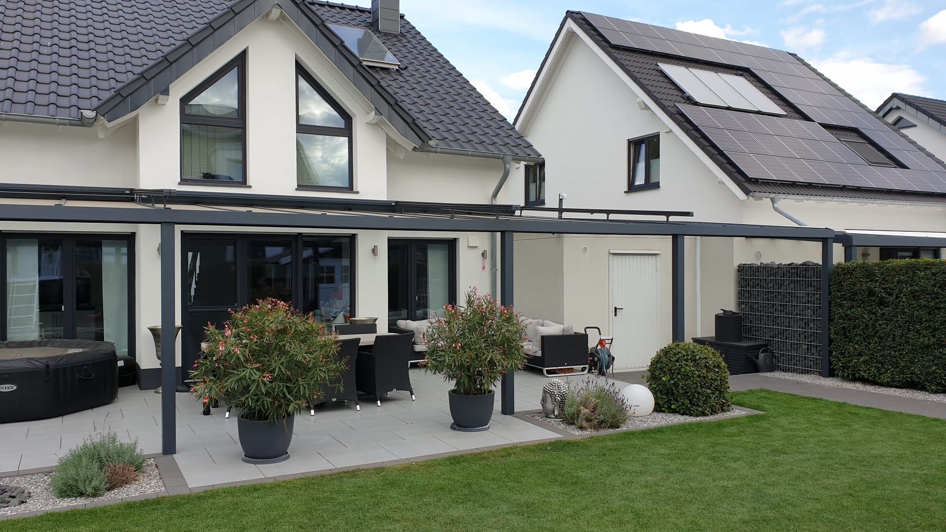 Eine Terrasse mit dunkelgrauer Pergola, Gartenmöbeln und Topfpflanzen vor zwei modernen Häusern an einem sonnigen Tag.