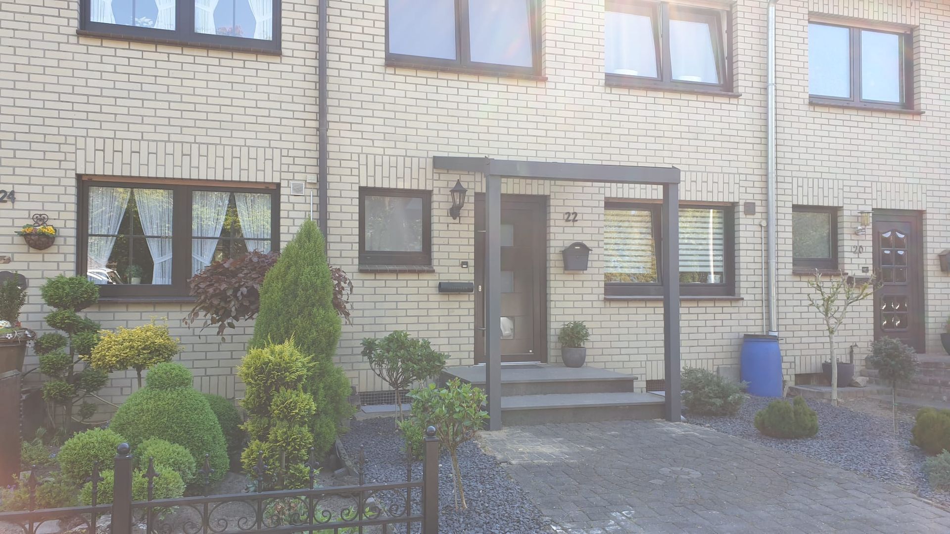 Ein helles Backsteinreihenhaus mit dunkel gerahmten Fenstern, einem überdachten Eingangsbereich und einem angelegten Vorgarten.