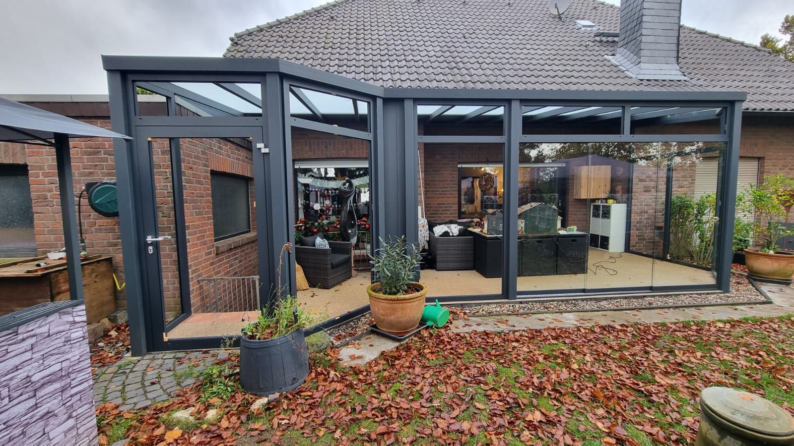 Ein moderner, dunkelgrauer Wintergarten mit Metallrahmen, der an die hintere Backsteinwand eines Hauses angebaut ist und sich zu einem Garten mit Laub öffnet.
