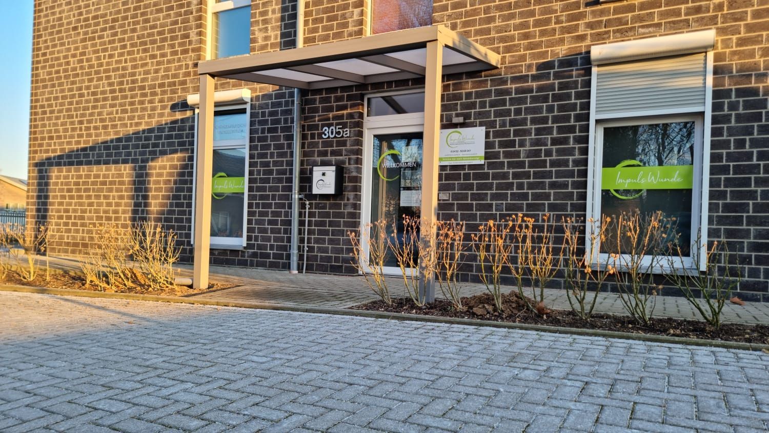 Der Eingang eines gemauerten Bürogebäudes mit kleinem Vordach, Hausnummer 205 und grüner Fensterbeschriftung unter sonnigem Himmel.