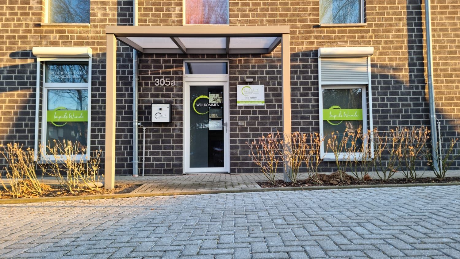 Der Eingang zu einem gemauerten Geschäftsgebäude mit grün gebrandeten Fenstern und einer Glastür unter einem kleinen Vordach.