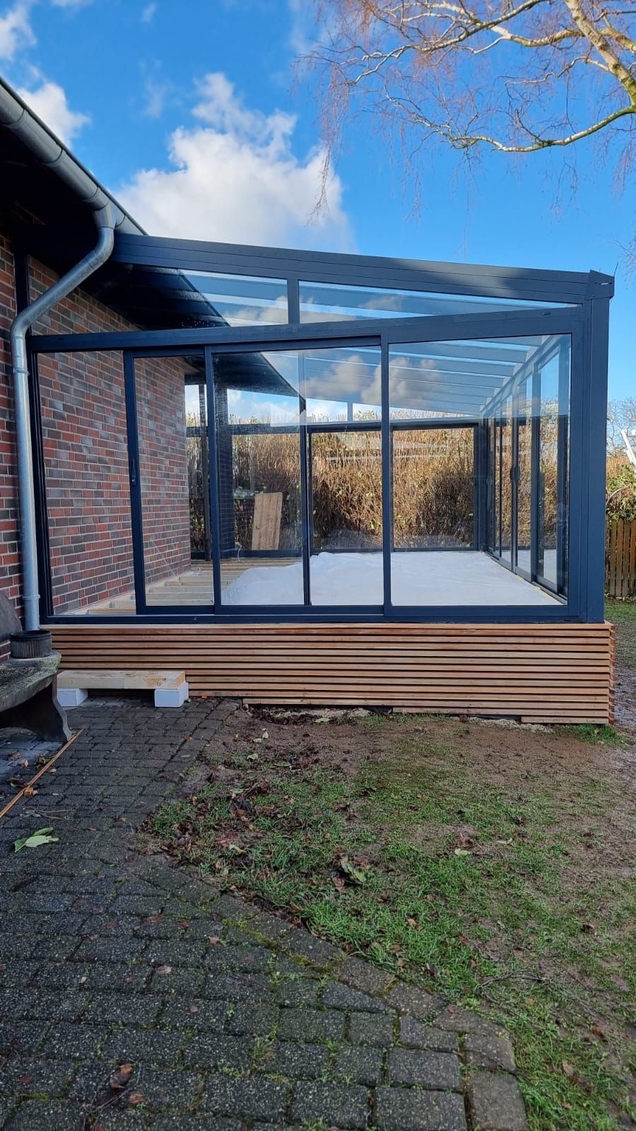 Ein moderner, dunkel gerahmter Glaswintergarten, der an ein Backsteinhaus angebaut ist und über Schiebetüren sowie eine Holzterrasse verfügt.
