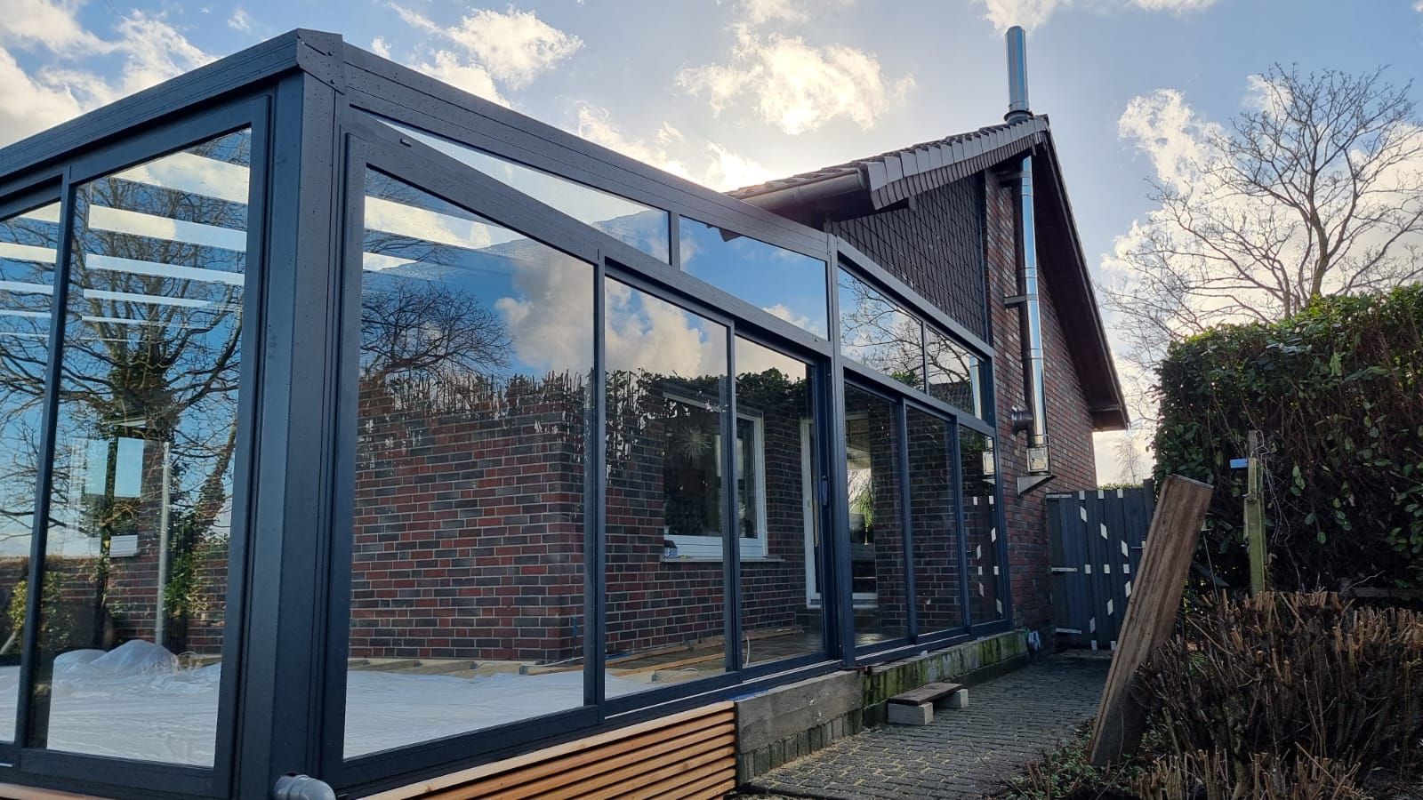 Ein schwarzgerahmter Glaswintergarten, der an ein Backsteinhaus angebaut ist, an einem sonnigen Tag mit Bäumen im Hintergrund.