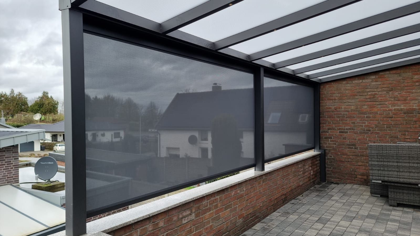 Ein schwarzer, einziehbarer Sichtschutz wurde unter einem modernen Terrassendach auf einer Ziegelterrasse mit Blick auf ein Wohngebiet installiert.