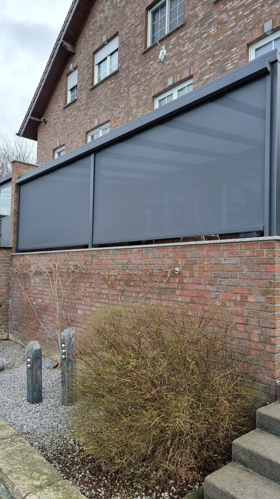 Ein dunkelgrauer Sichtschutz oder eine ausziehbare Jalousie, die an einer gemauerten Terrassenkonstruktion neben einem Haus angebracht ist.