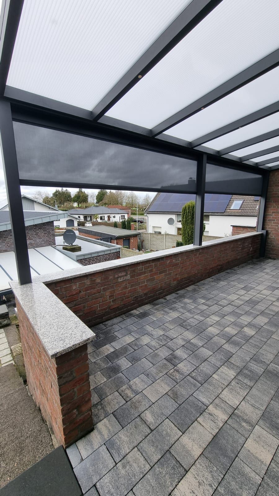 Eine Terrasse mit grauen Steinplatten, eine halbhohe Ziegelmauer mit Granitabdeckung und eine moderne schwarze Metallpergola mit Sonnenschutz.