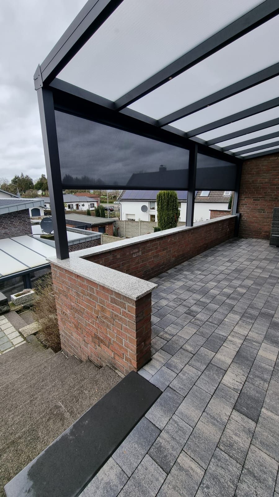 Eine Terrasse mit einer dunkelgrauen Metallpergola, einer halbhohen Backsteinmauer und einem grauen Pflasterboden mit Blick auf Vorstadthäuser.
