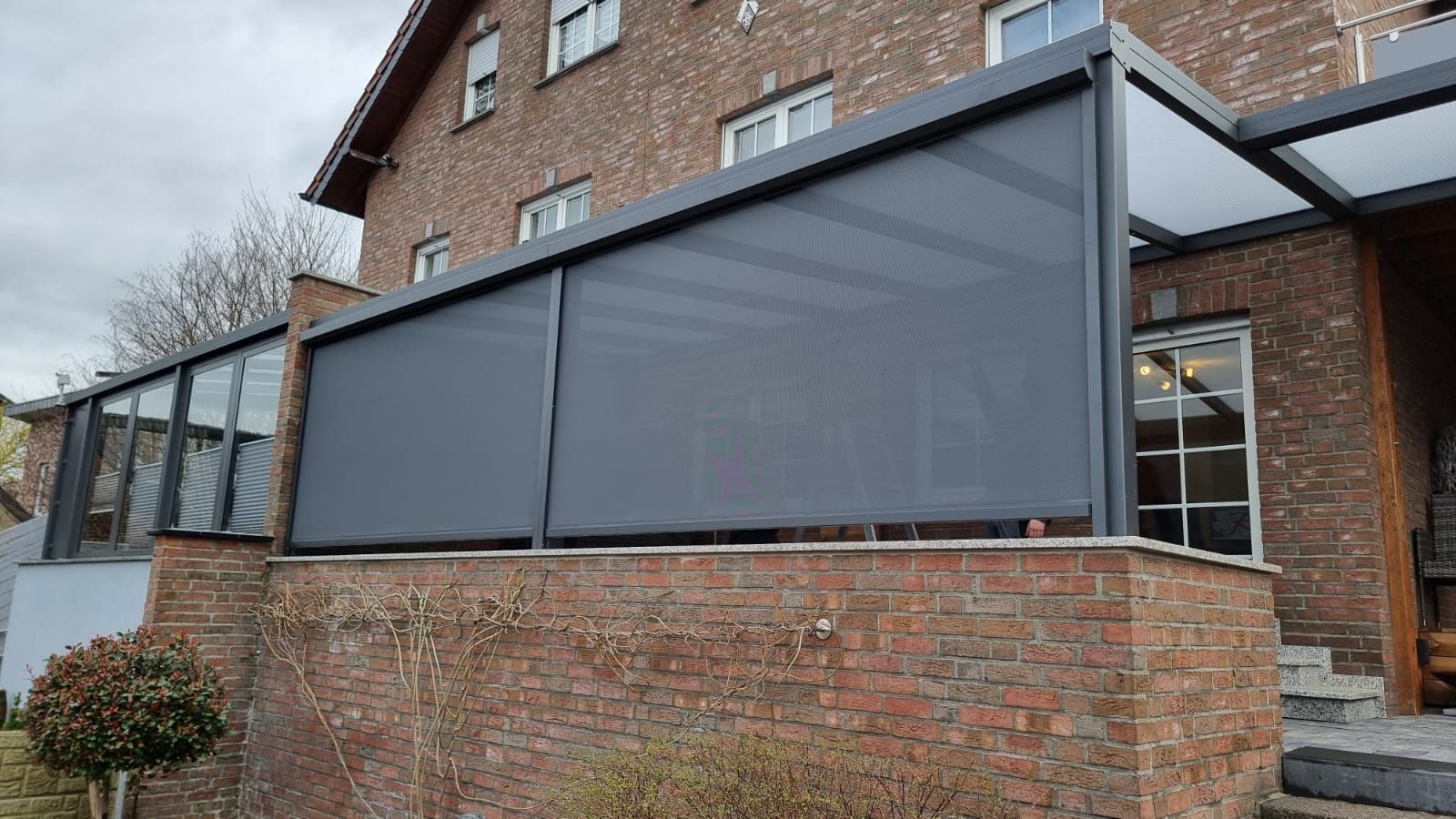 Ein Backsteinhaus mit einer modernen Terrassenüberdachung, die mit dunkelgrauen, vertikalen Sichtschutzwänden an der Außenwand ausgestattet ist.