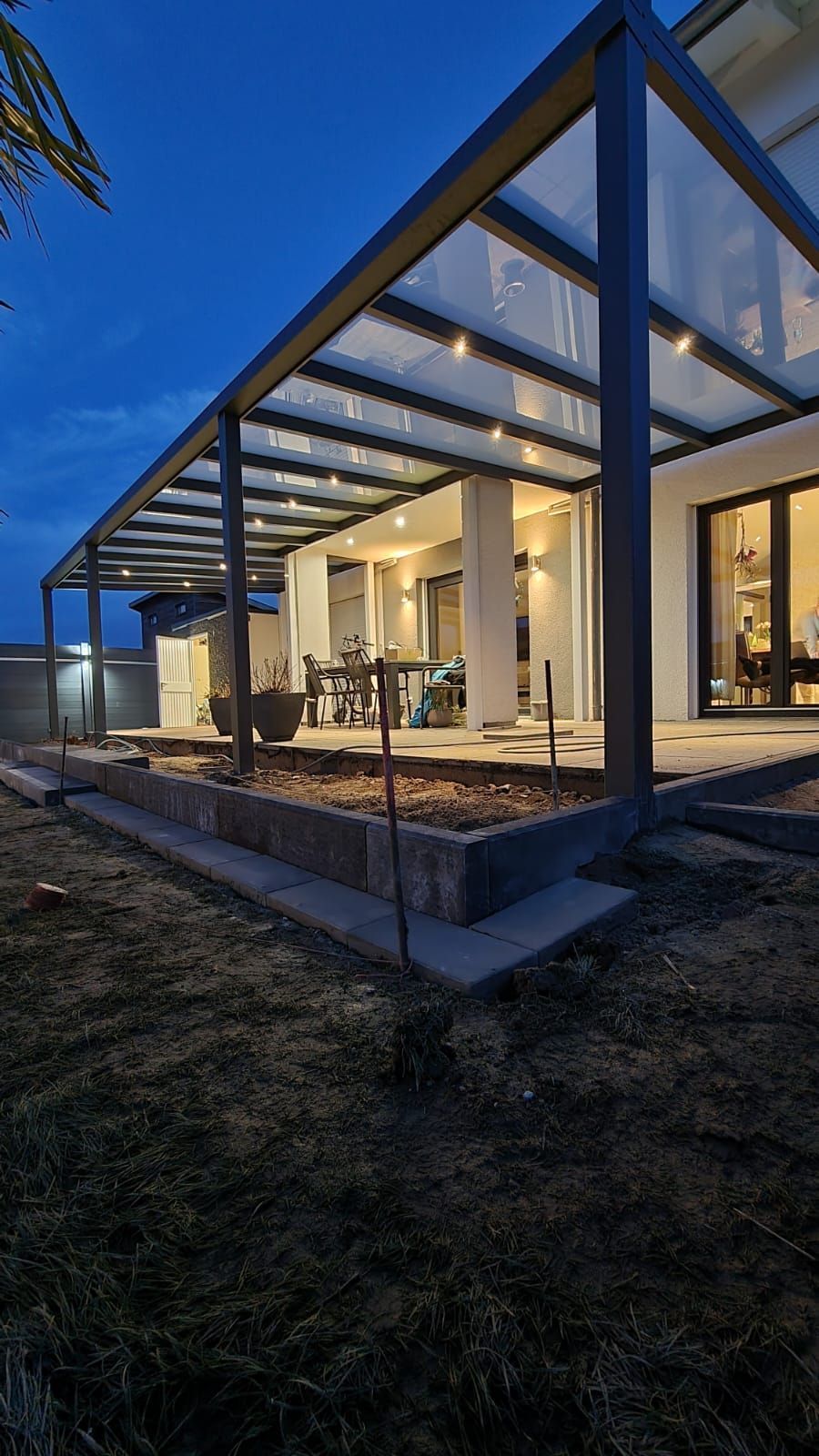 Moderne Terrassenkonstruktion mit Glasdach, dunklem Metallrahmen und integrierter Dämmerungsbeleuchtung.