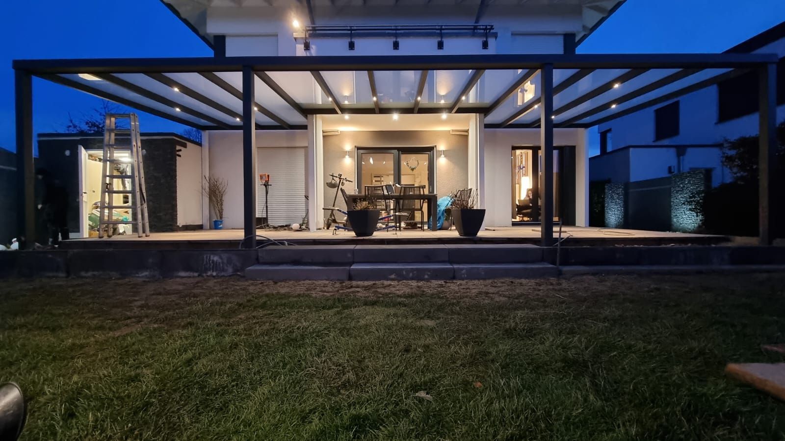 Eine beleuchtete, moderne Terrasse mit überdachter Pergola in der Abenddämmerung, mit Gartenmöbeln und einer Treppe, die zu einem Rasen führt.
