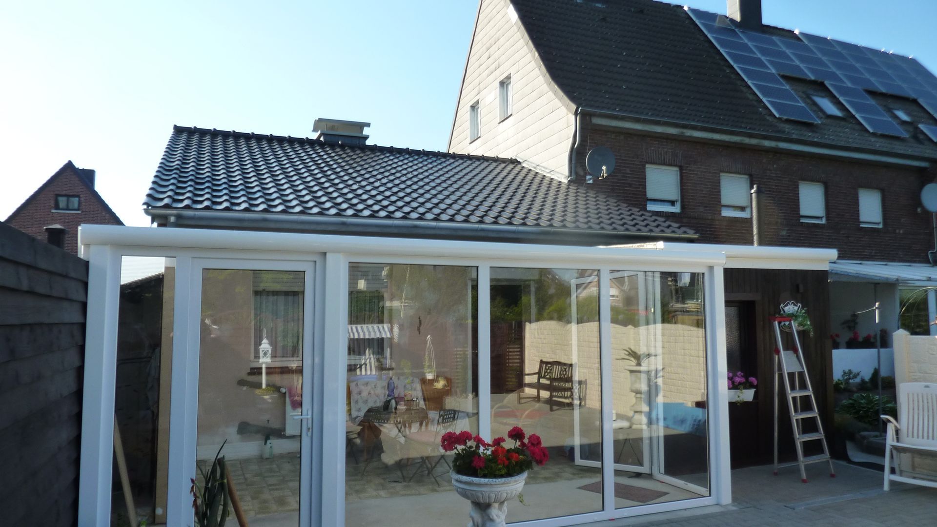Neben einem Backsteinhaus mit Solarpaneelen auf dem Dach befindet sich ein verglaster Terrassenanbau mit weißem Rahmen.