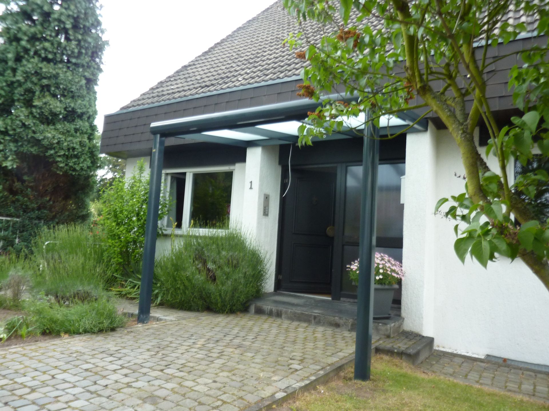 Ein weißer Hauseingang mit schwarzer Tür, einem Glasvordach und einem Kopfsteinpflasterweg, aus einer leicht schrägen Perspektive betrachtet.