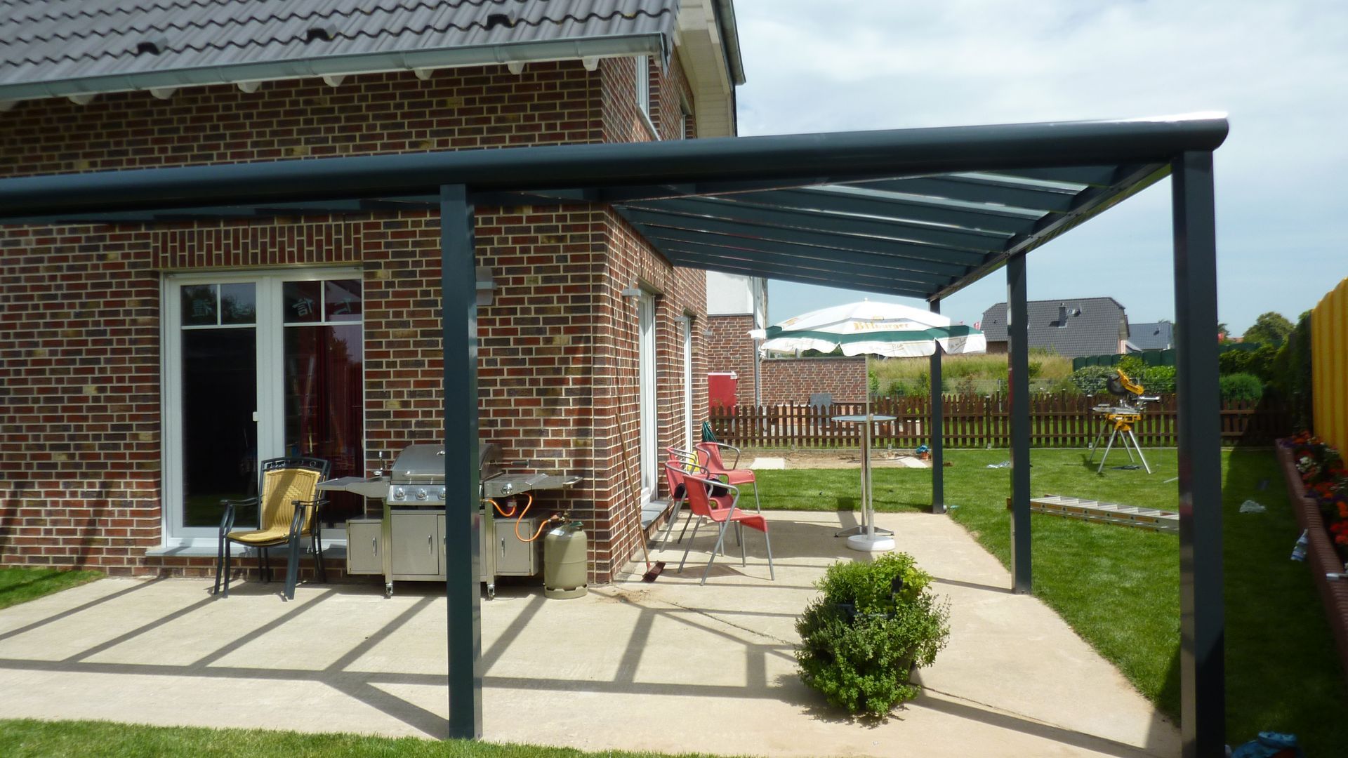 Von einem Backsteinhaus erstreckt sich eine dunkel umrahmte Terrassenüberdachung über einen gepflasterten Bereich mit Grill, Sitzgelegenheiten im Freien und einem kleinen Busch.