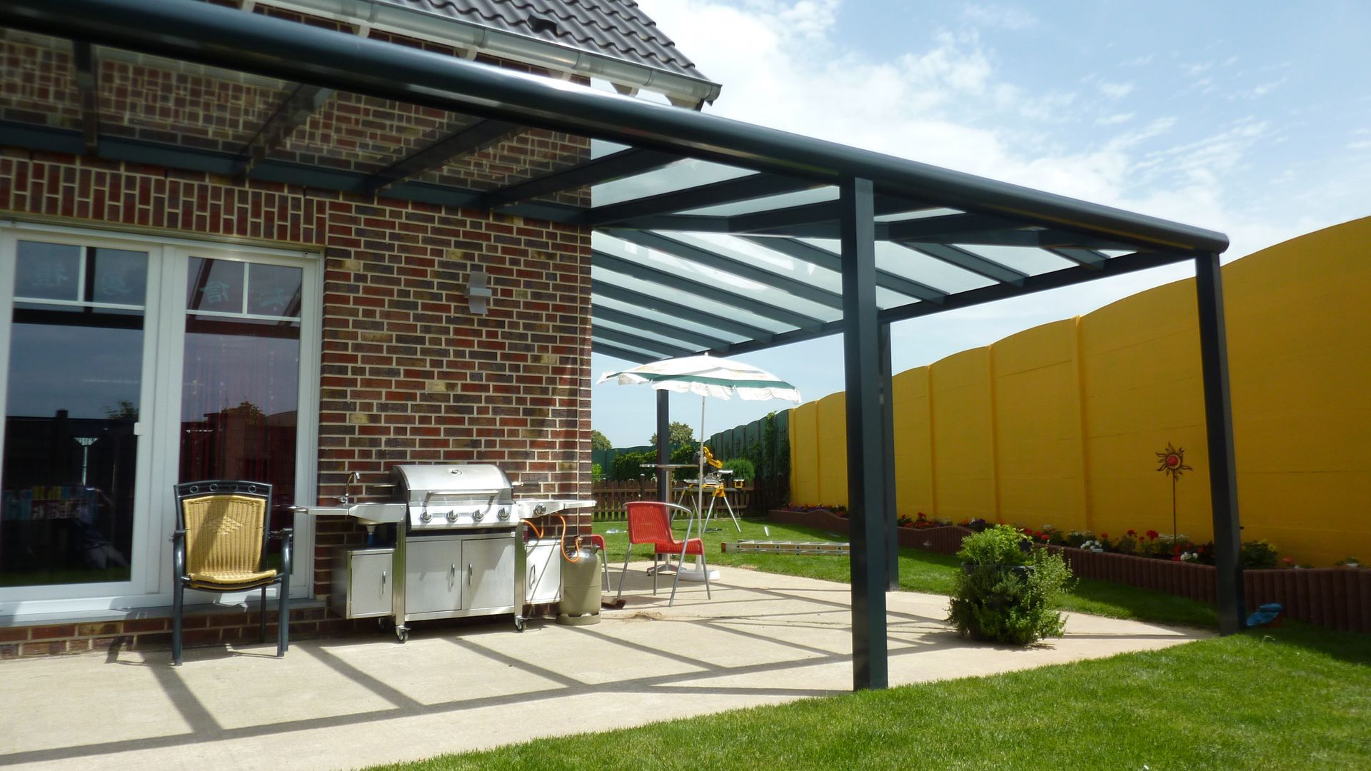 Eine Terrasse mit einer Pergola unter Glasdach, einem Edelstahlgrill und Gartenmöbeln vor einem Backsteinhaus und einer gelben Mauer.