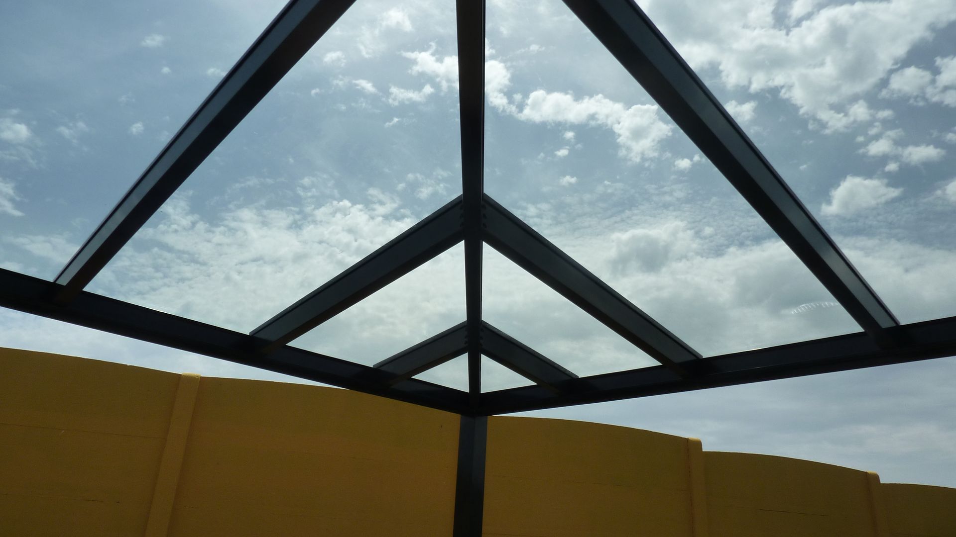 Eine Ansicht aus der Froschperspektive auf ein schwarzes Metallgerüst eines Pavillons vor blauem Himmel mit weißen Wolken und einer gelben Mauer im Hintergrund.