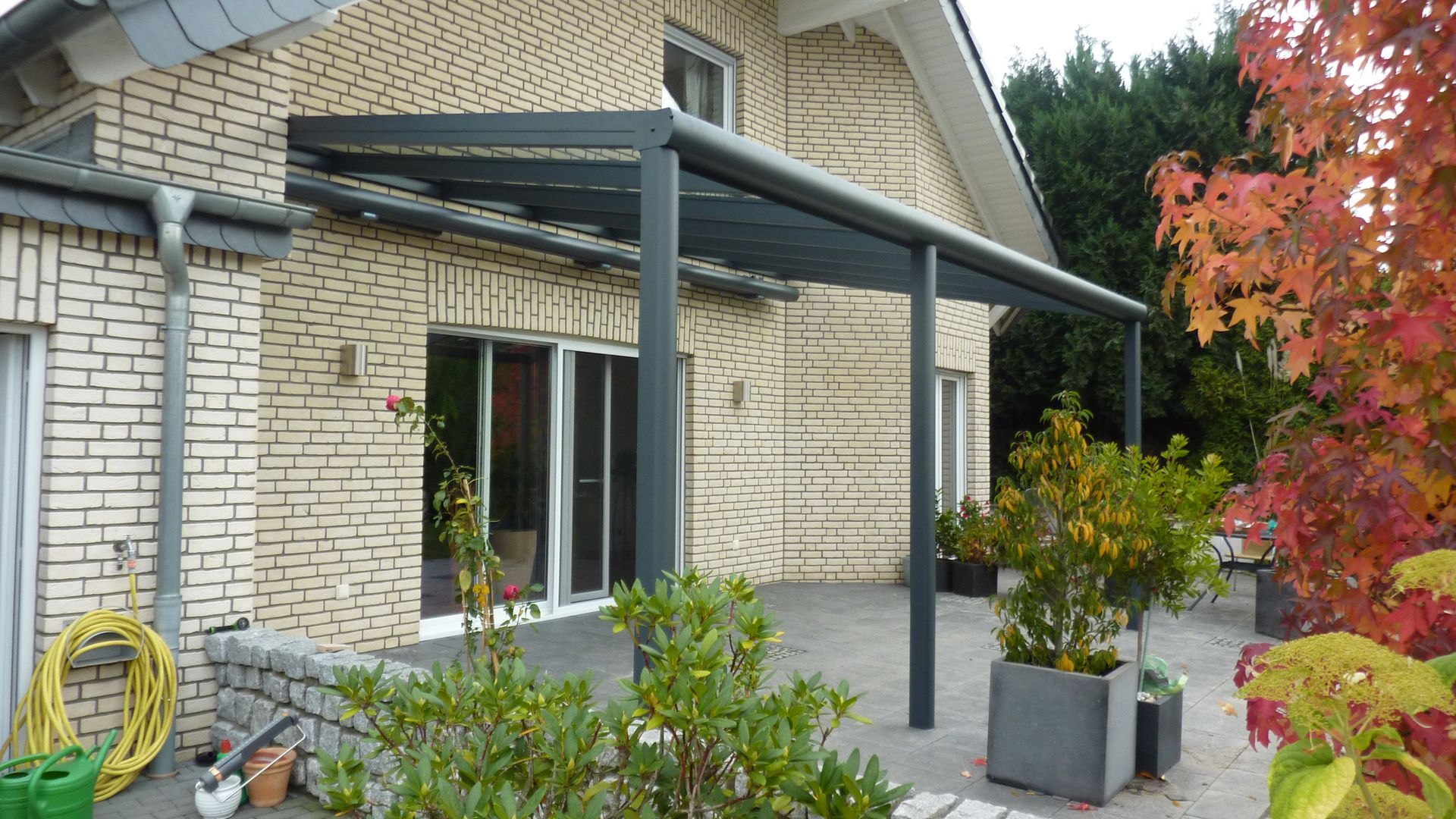 Eine graue, moderne Terrassenüberdachung an einem hellen Backsteinhaus mit Blick auf eine gepflasterte Terrasse mit Topfpflanzen.