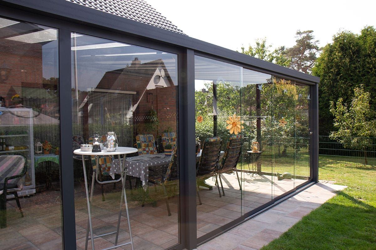 Eine verglaste Terrasse mit dunklen Metallrahmen, auf der ein Tisch und Stühle stehen, vor einem Haus und einem Rasengarten.
