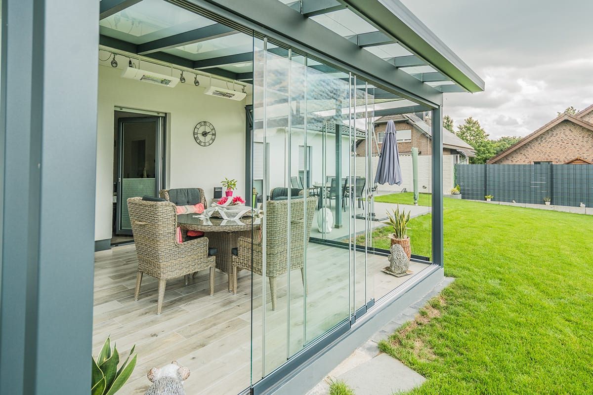 Eine verglaste Terrasse mit Korbmöbeln, einem Tisch und Blick auf einen grasbewachsenen Hinterhof unter bedecktem Himmel.