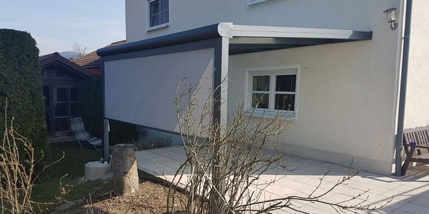 Eine Terrasse mit einem grauen, einziehbaren Sichtschutz, der an einem dunklen Rahmen an der Seite eines weißen Hauses befestigt ist.