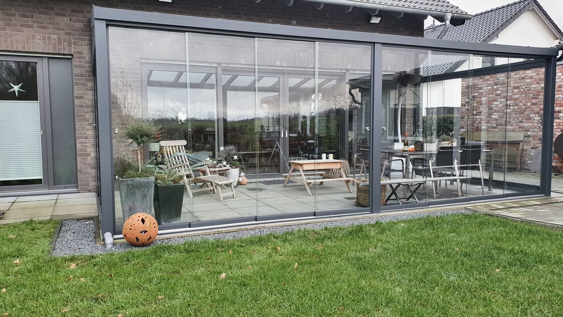 Eine moderne, verglaste Terrasse mit dunklem Rahmen, die an ein Backsteinhaus angebaut ist und auf einen grünen Rasen mit einem kleinen orangefarbenen Ball blickt.