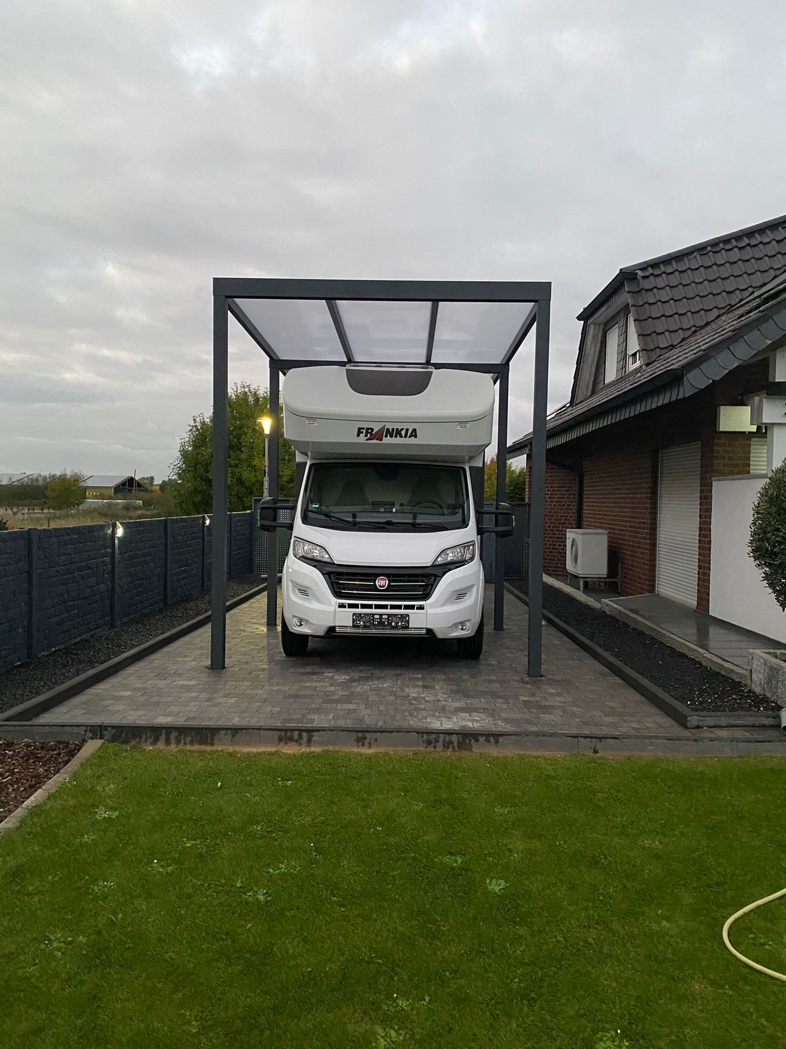 Ein weißes Wohnmobil parkt unter einem modernen, grauen Metallcarport neben einem Backsteinhaus mit Rasen im Vordergrund.