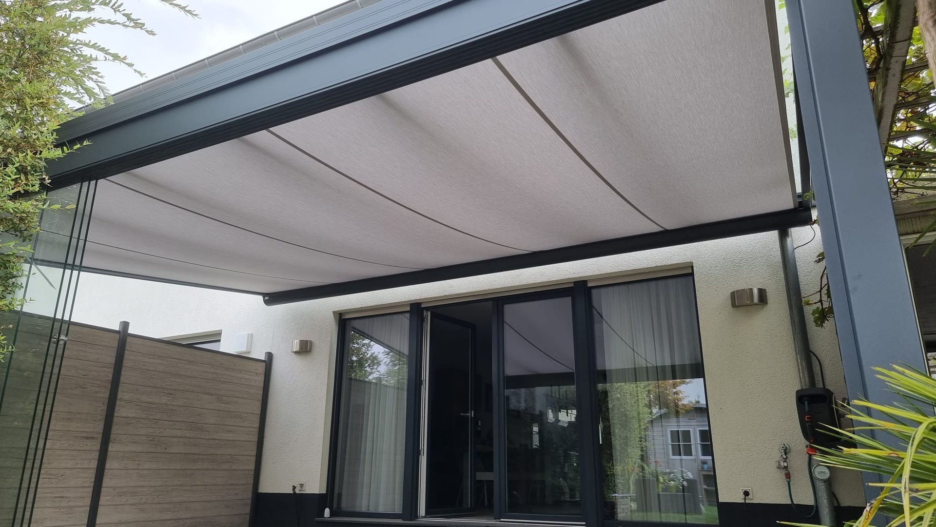 Eine moderne Terrassenkonstruktion mit einem einziehbaren weißen Vordach, die an ein helles Gebäude mit Glasschiebetüren angebaut ist.