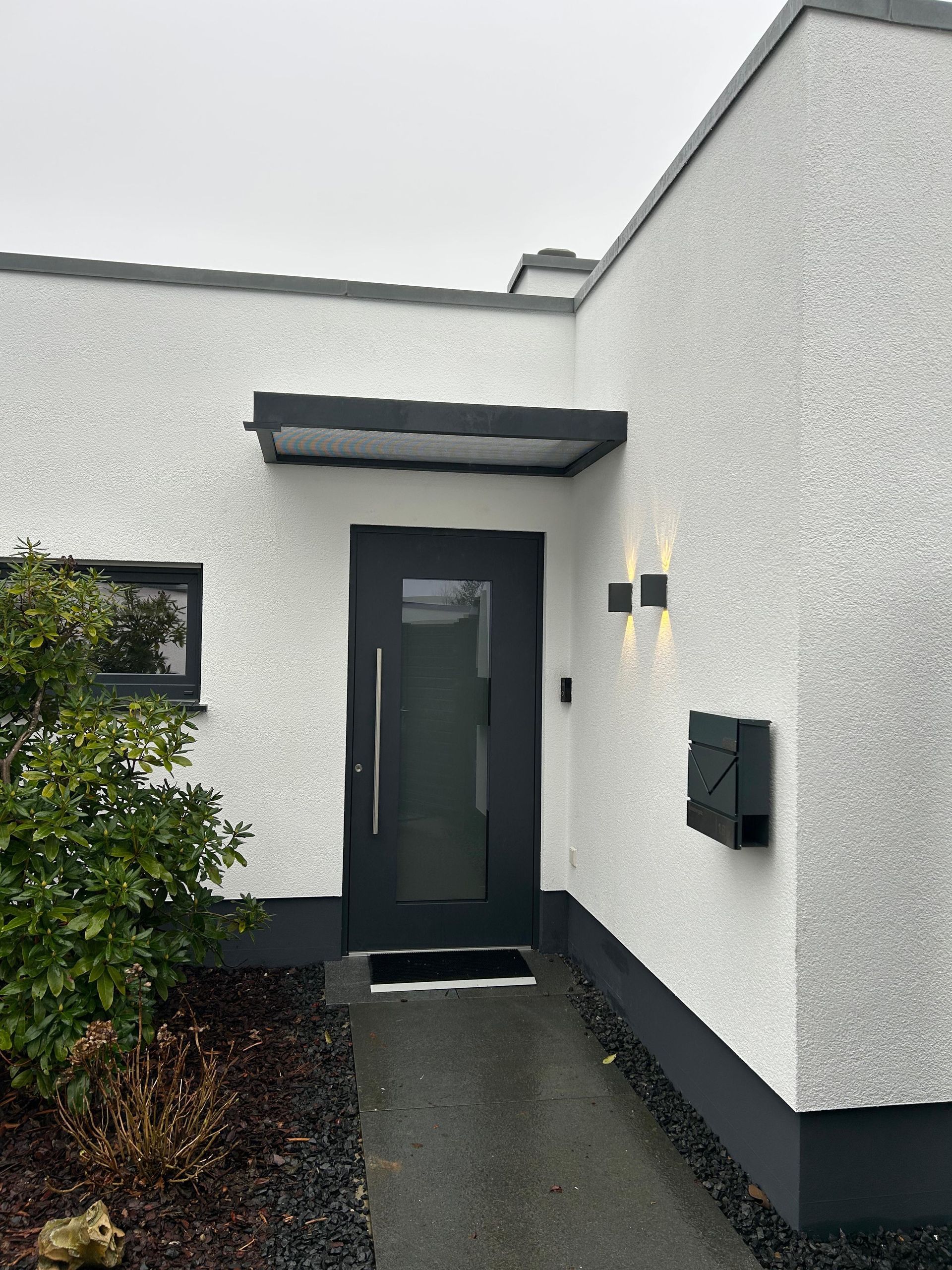 Moderner Eingang mit dunkelgrauer Tür, kleinem, flachem Vordach, Außenwandleuchten und einem Briefkasten an einer weißen, strukturierten Wand.